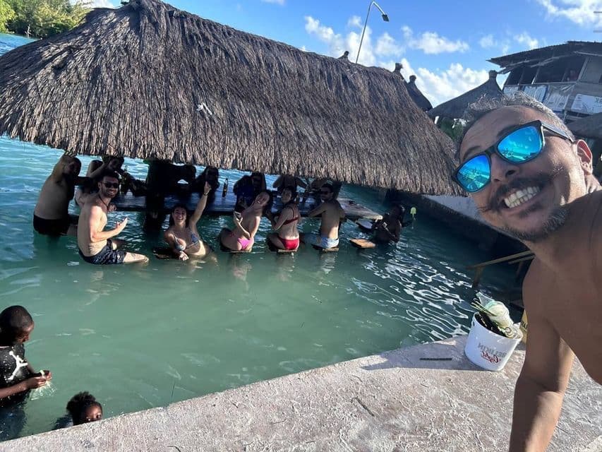 Un uomo si scatta un selfie sorridente mentre il suo gruppo WeRoad si rilassa a un bar galleggiante con tetto in paglia, immerso in acque cristalline.