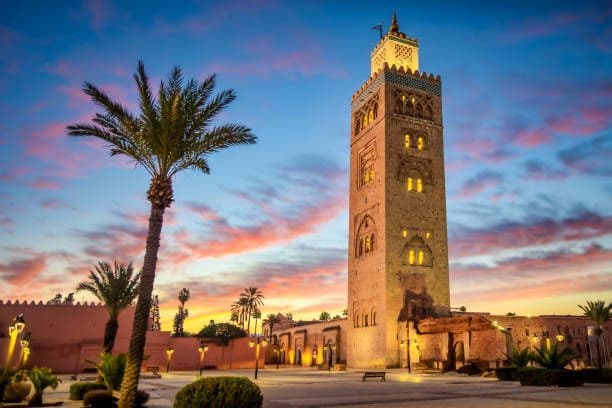 Un alto minareto di pietra è illuminato in un cortile con una palma, sullo sfondo di un cielo colorato al tramonto.
