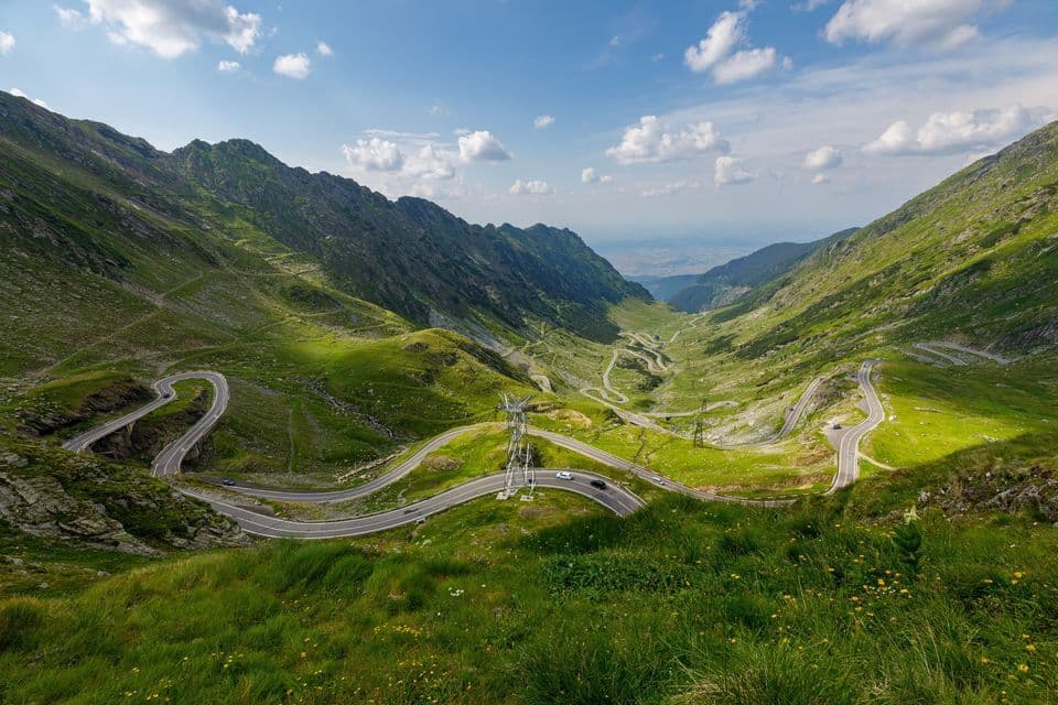 Una carretera asfaltada serpenteante con coches atraviesa un valle de montaña de un verde vibrante bajo un cielo azul con nubes blancas.