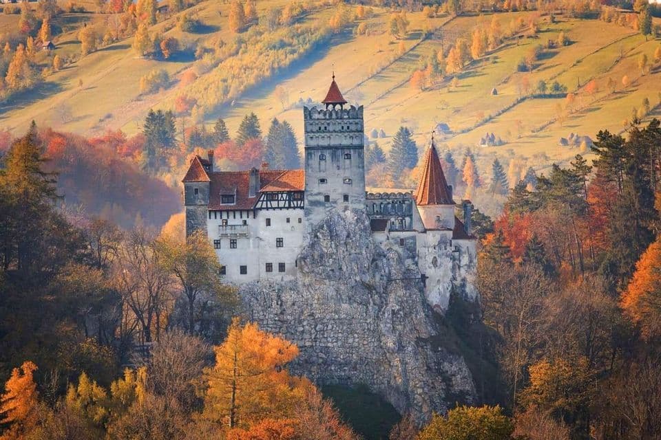 Un gran castillo de tejados rojos se alza sobre un acantilado rocoso, rodeado por un bosque de árboles otoñales coloridos y colinas ondulantes.