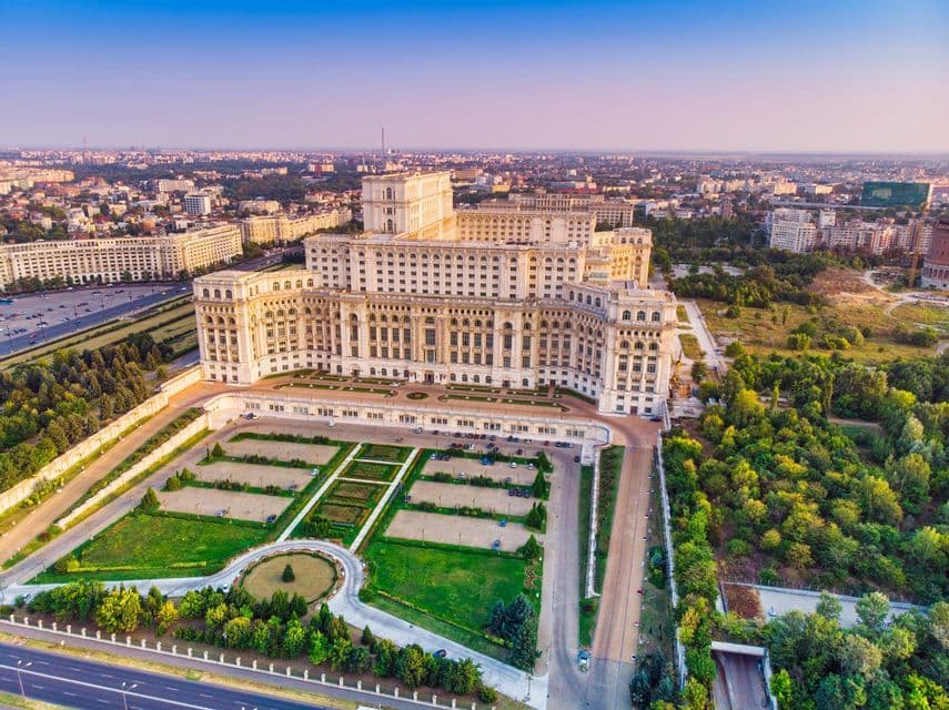 Una vista aérea de un gran y ornamentado edificio gubernamental rodeado de céspedes bien cuidados, carreteras y un vasto paisaje urbano al atardecer.