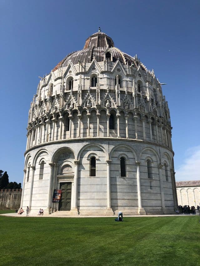 Ein großes, rundes, weißes Steingebäude mit verzierten Bögen und einer Kuppel, gesehen über eine weite grüne Wiese hinweg unter einem klaren blauen Himmel.