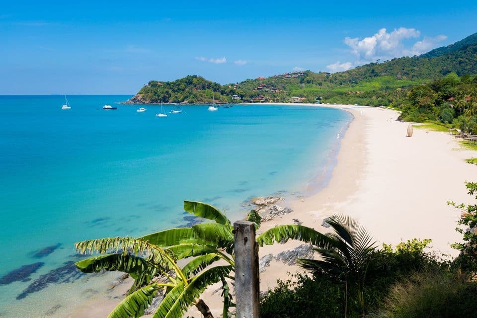 A high-angle view of a tropical bay with a white sand beach, clear turquoise water, and sailboats anchored offshore.