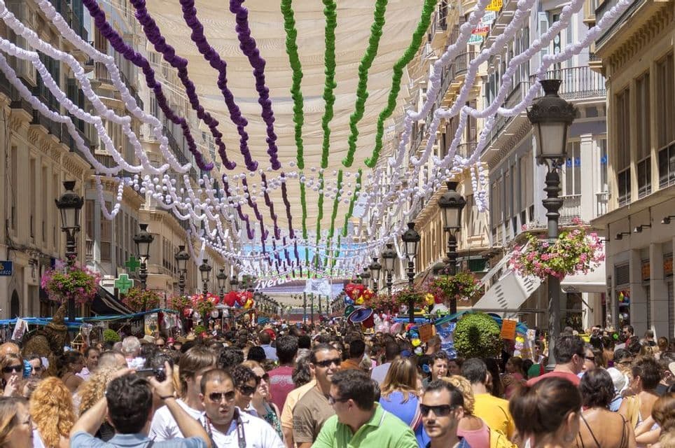 Una grande folla di persone percorre una strada stretta, decorata con ghirlande colorate sospese e un baldacchino bianco, durante un festival.