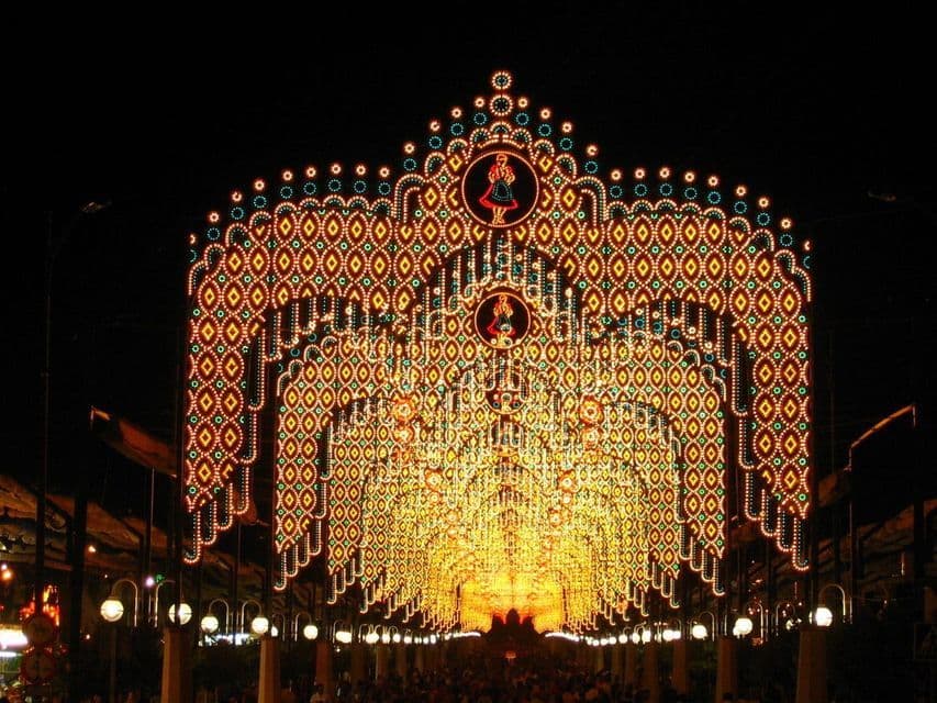 Un arco ornato di luci gialle e verdi crea un tunnel sopra una strada affollata durante un festival notturno.