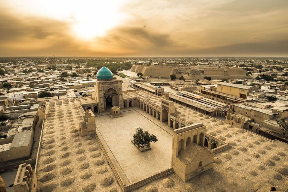 An aerial view of a historic city at sunset, featuring a large mosque with a turquoise dome and a central courtyard.