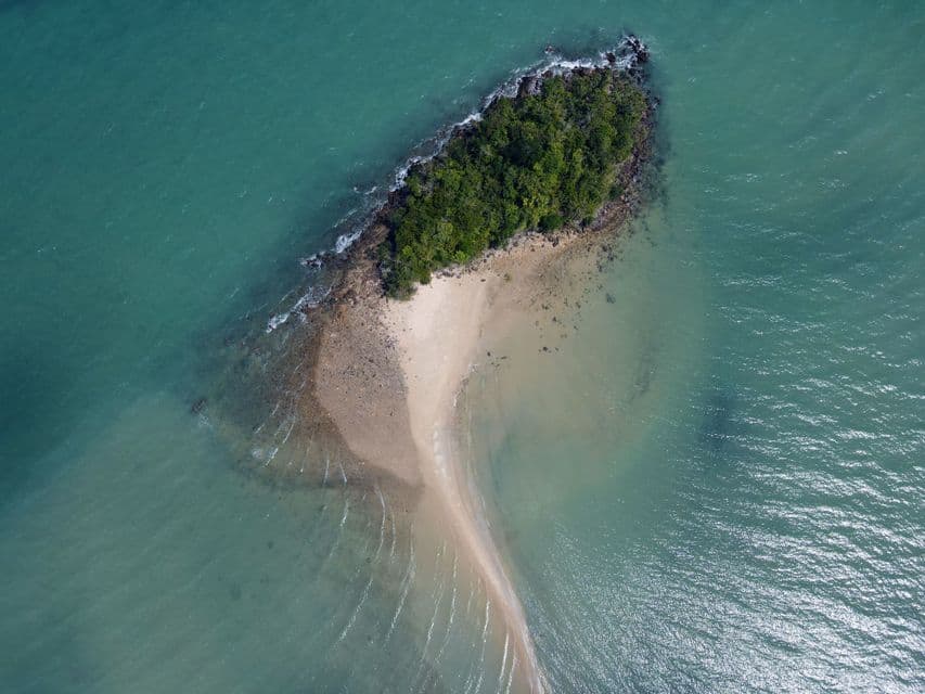 An aerial top-down view of a small, tree-covered island with a long sandbar extending into the turquoise sea.