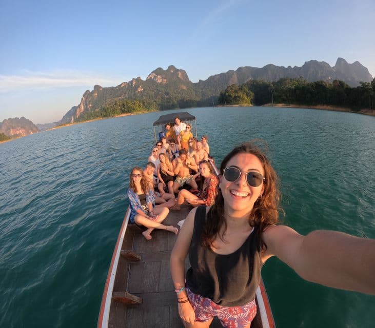 A woman takes a selfie with a WeRoad group trip on a long-tail boat on a lake surrounded by mountains.