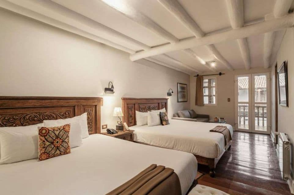 Ein Hotelzimmer mit zwei Doppelbetten, geschnitzten Holz-Kopfteilen, einem dunklen Holzfußboden und einer weißen Balkendecke.