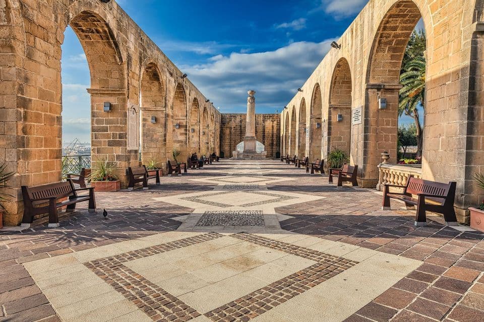 A long stone-paved walkway flanked by large arches, with wooden benches lining the path towards a distant monument.