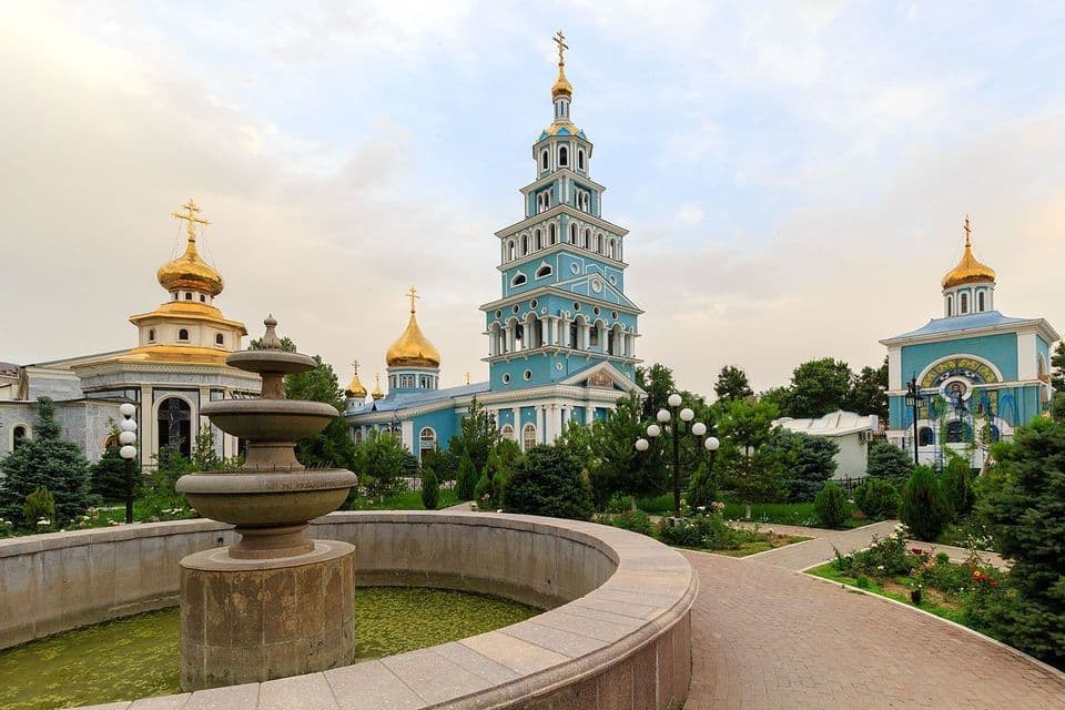 Mehrere blaue orthodoxe Kirchen mit goldenen Zwiebeltürmen stehen in einem Garten mit einem steinernen Brunnen im Vordergrund.