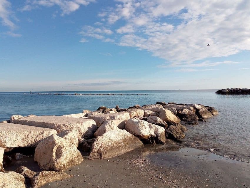 Una scogliera frangiflutti si estende da una spiaggia sabbiosa nel mare calmo, sotto un cielo azzurro con nuvole bianche.