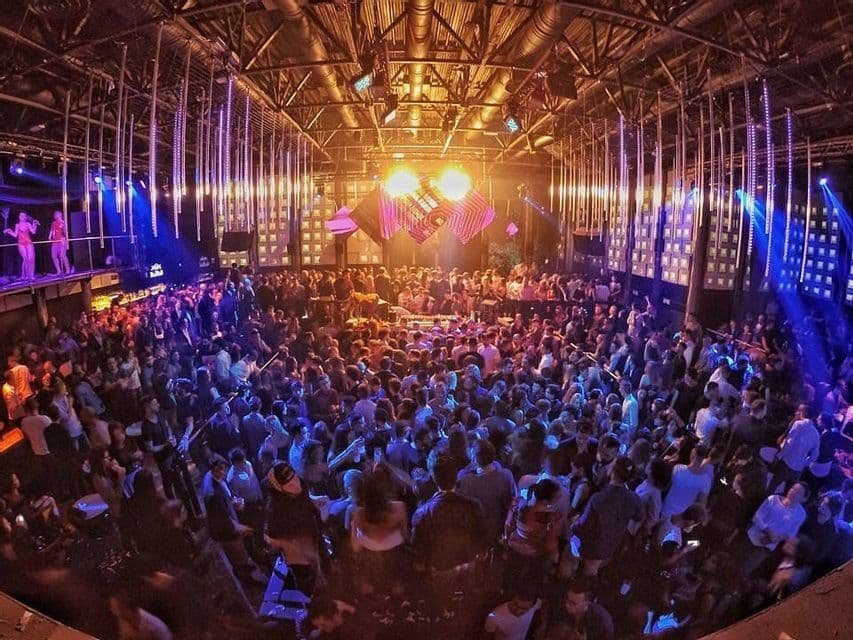 Una veduta grandangolare di una grande folla che balla in una discoteca sotto luci di scena colorate e installazioni luminose sospese.