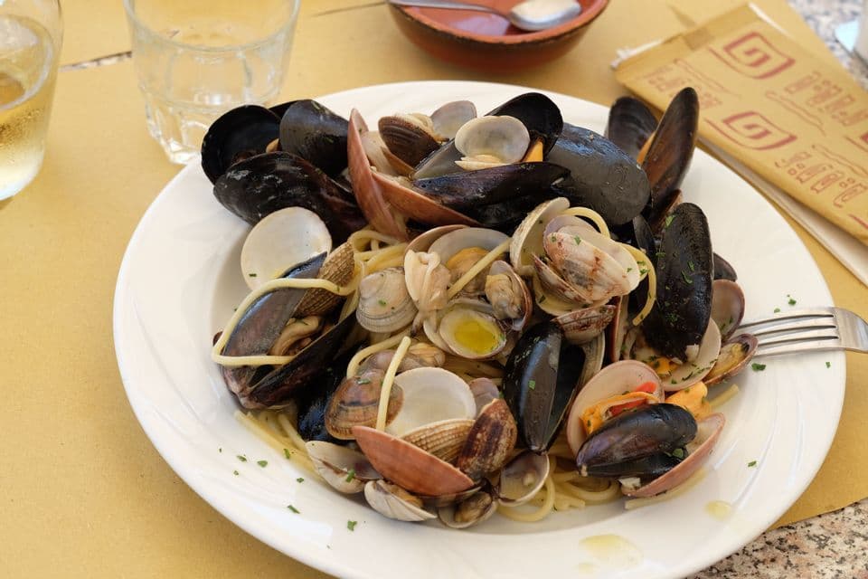 Un piatto bianco di spaghetti mescolati con cozze e vongole, poggiato su una tovaglietta di carta gialla su un tavolo.