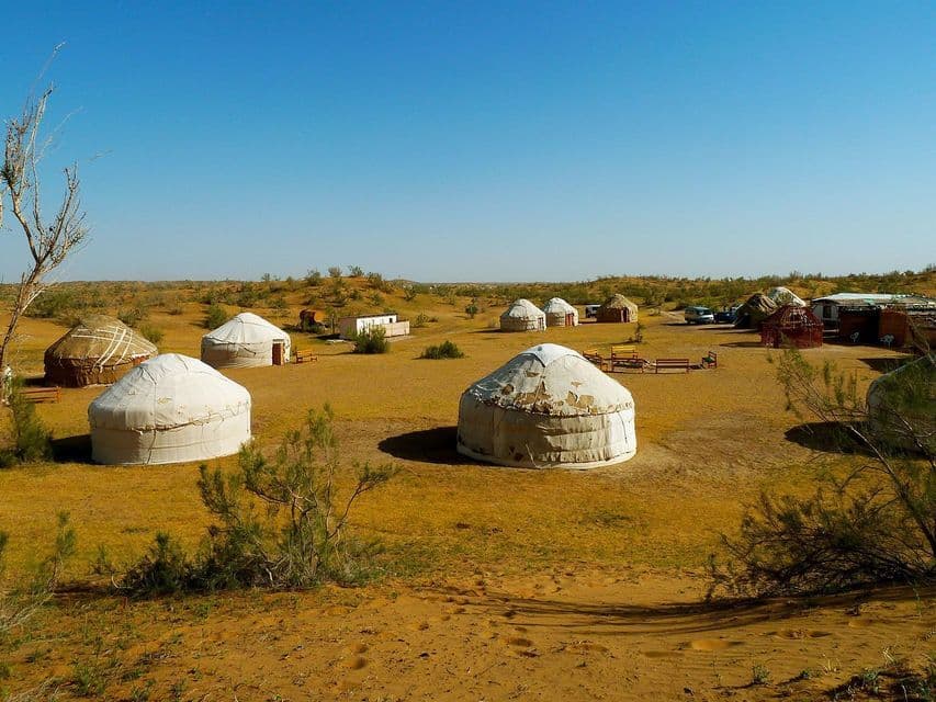 Un campamento de yurtas tradicionales dispersas por un paisaje árido y arenoso con arbustos bajos y verdes bajo un cielo azul claro.