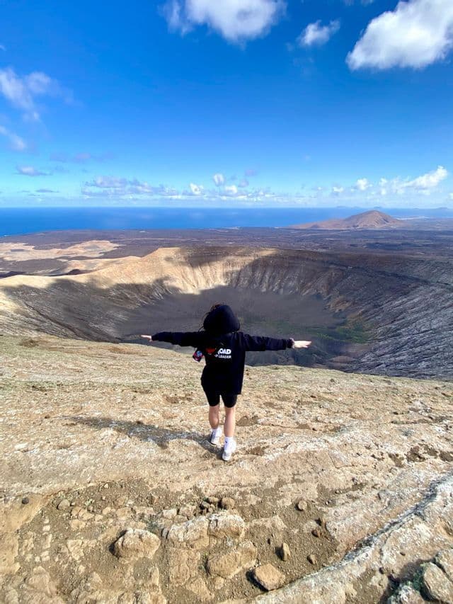 Una persona con una felpa nera con cappuccio si trova sull'orlo di un grande cratere vulcanico, ammirando un vasto paesaggio e l'oceano.