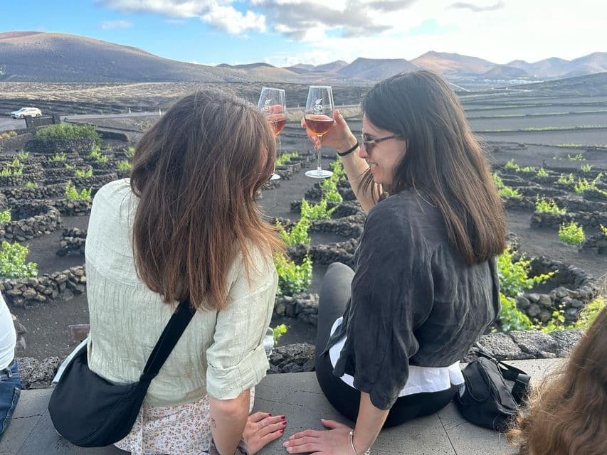 Due donne di un viaggio di gruppo WeRoad brindano con calici di vino rosé, con vista su un vigneto vulcanico e montagne sullo sfondo.