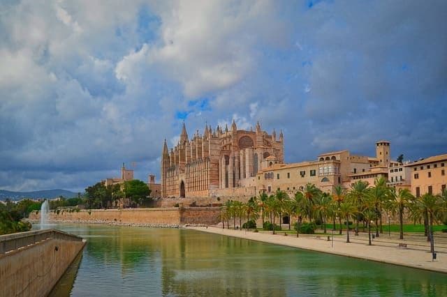 Una gran catedral gótica y edificios adyacentes se alzan junto a un cuerpo de agua bordeado de palmeras bajo un cielo nublado.
