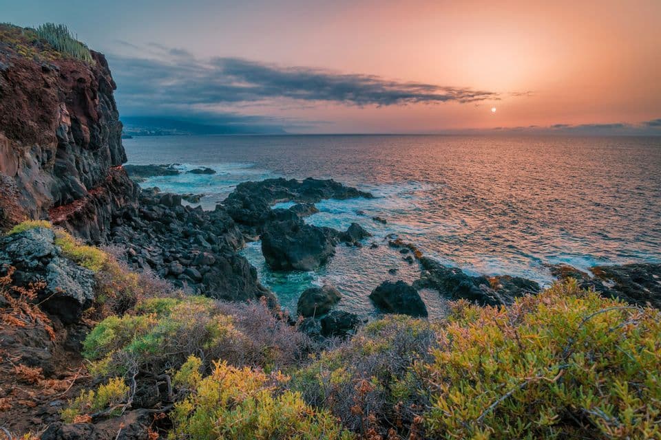 Una costa rocciosa con arbusti si affaccia sull'oceano mentre il sole tramonta all'orizzonte sotto un cielo nuvoloso.