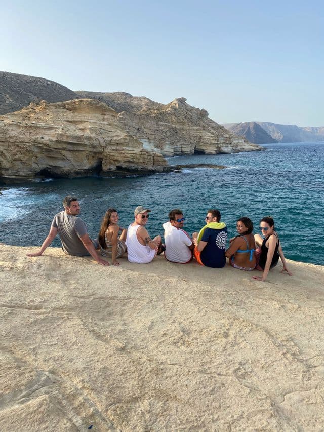 Un viaje en grupo de WeRoad sentados juntos en un acantilado rocoso, observando el océano y formaciones rocosas estratificadas a lo largo de la costa.