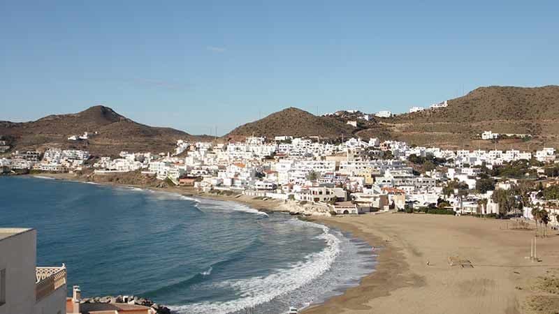 Un pueblo costero de edificios blancos se asienta a lo largo de una amplia playa de arena, con olas suaves y colinas áridas al fondo bajo un cielo despejado.