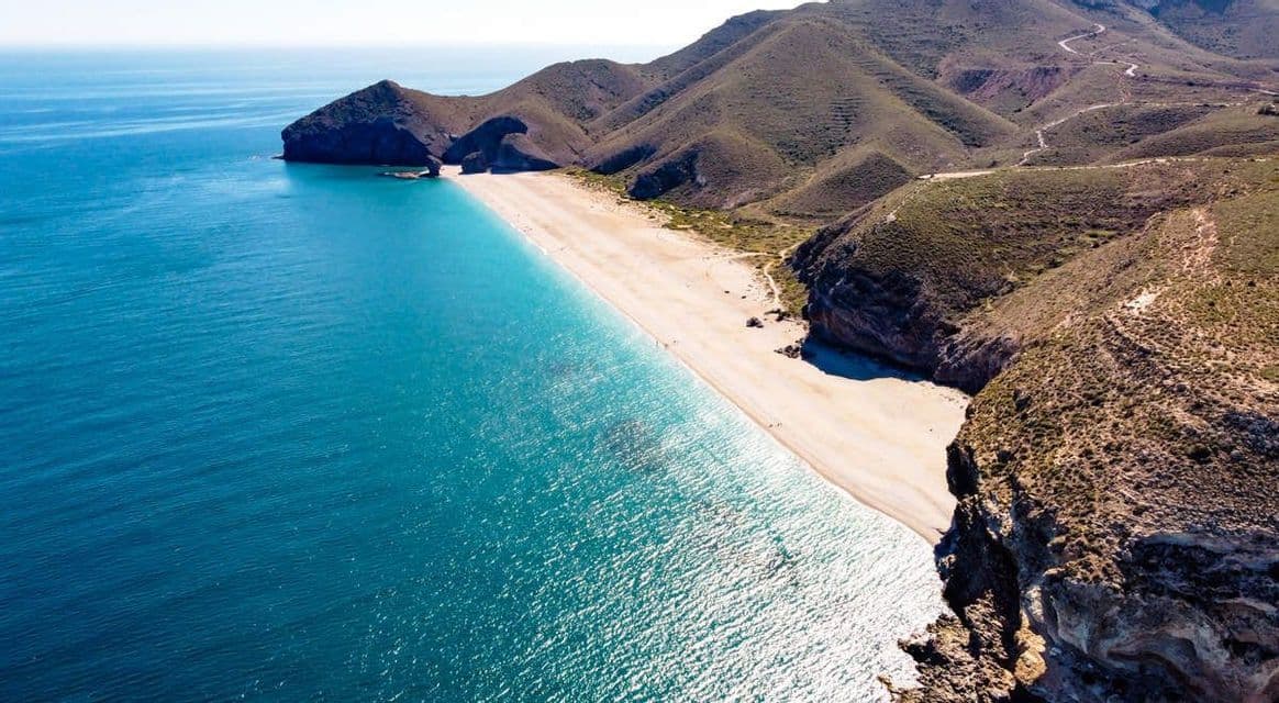 Una vista aérea muestra una larga playa de arena situada entre las aguas turquesas del océano y colinas áridas y ondulantes bajo un cielo despejado.