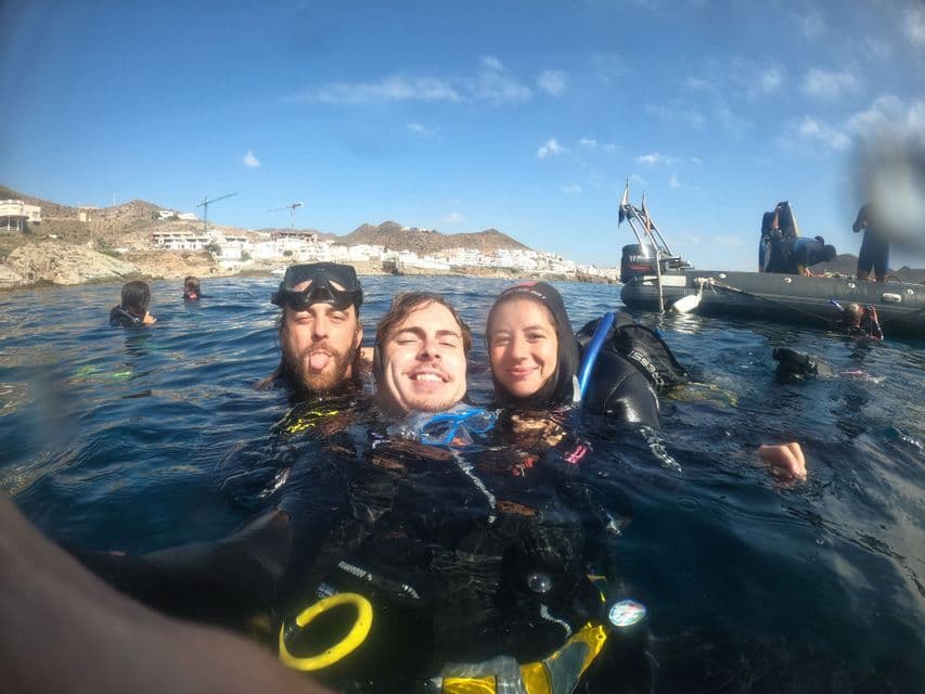 Tres personas en un viaje en grupo de WeRoad se toman una selfie en el mar usando trajes de neopreno y equipo de buceo, con un barco y la costa detrás de ellos.