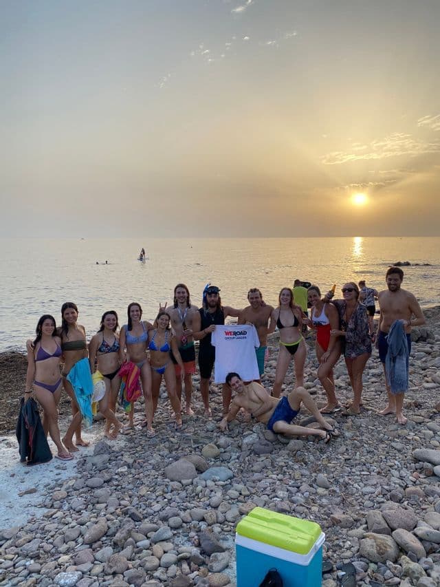 Un grupo de WeRoad posa en traje de baño en una playa rocosa al atardecer, sosteniendo una camiseta de WeRoad.