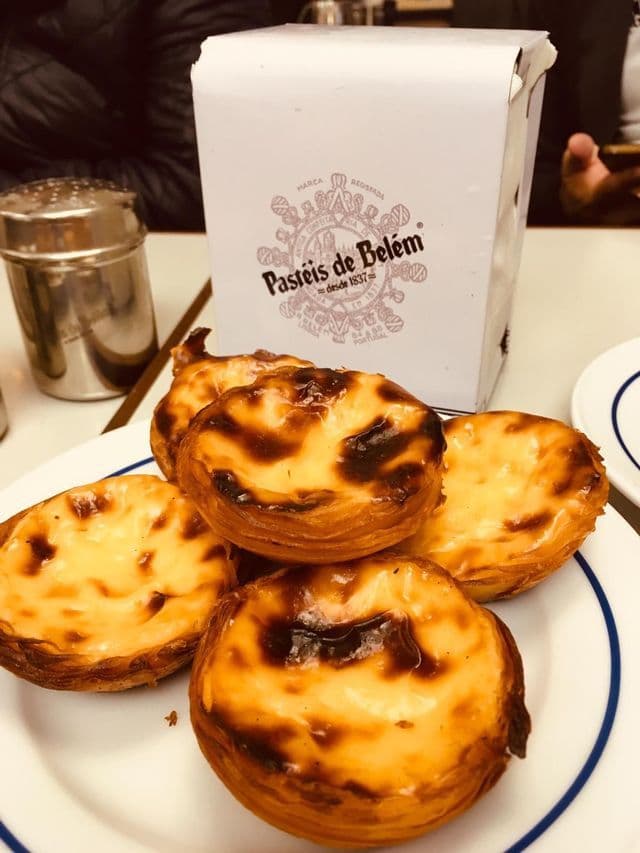 A close-up of several Pastéis de Belém custard tarts with caramelized tops on a plate, next to their branded box.