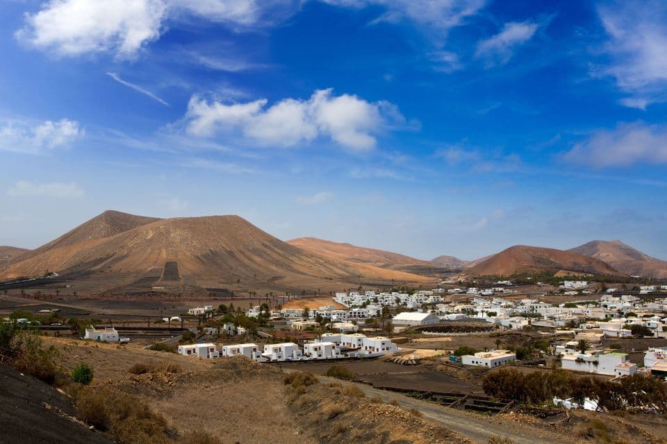 Un borgo di case bianche incastonato in un paesaggio vulcanico con colline brune sotto un cielo azzurro e nuvole sparse.