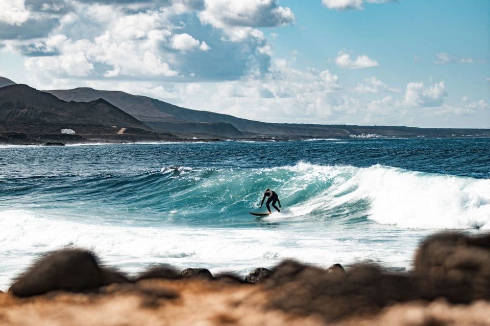 Un surfista in muta nera cavalca un'onda turchese, con una costa rocciosa e montuosa sullo sfondo.