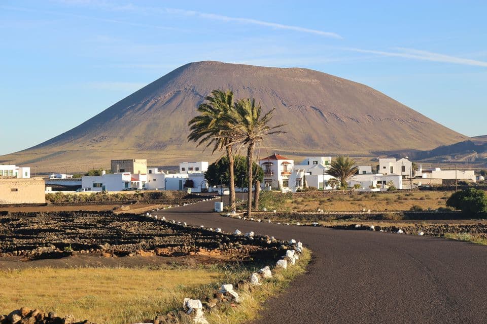 Una strada tortuosa attraversa un paesaggio vulcanico verso un piccolo villaggio di edifici bianchi adagiato ai piedi di un grande vulcano.