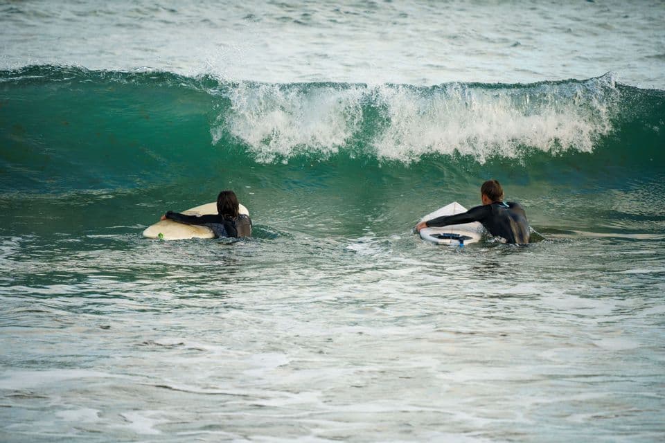 Due persone in muta galleggiano sulle loro tavole da surf nell'oceano mentre un'onda si infrange dietro di loro.