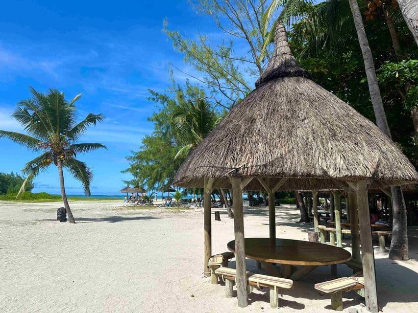 Un gazebo con tetto di paglia, con un tavolo e panche di legno, si trova su una spiaggia di sabbia bianca, circondato da palme sotto un cielo azzurro e limpido.