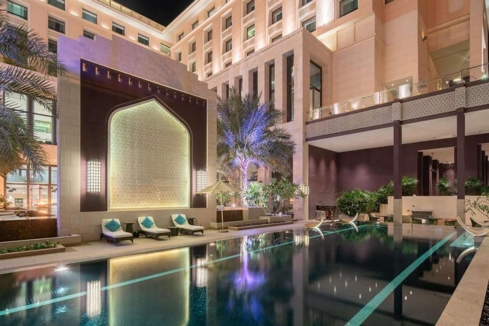 Une piscine extérieure dans la cour d'un hôtel la nuit, avec une architecture illuminée et des chaises longues se reflétant sur l'eau calme.