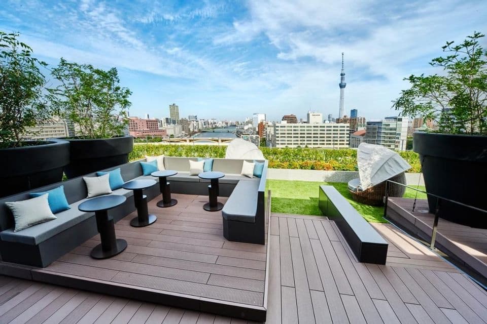 Une terrasse moderne sur le toit avec des canapés et tables gris, offrant une vue sur un paysage urbain avec une haute tour sous un ciel bleu.