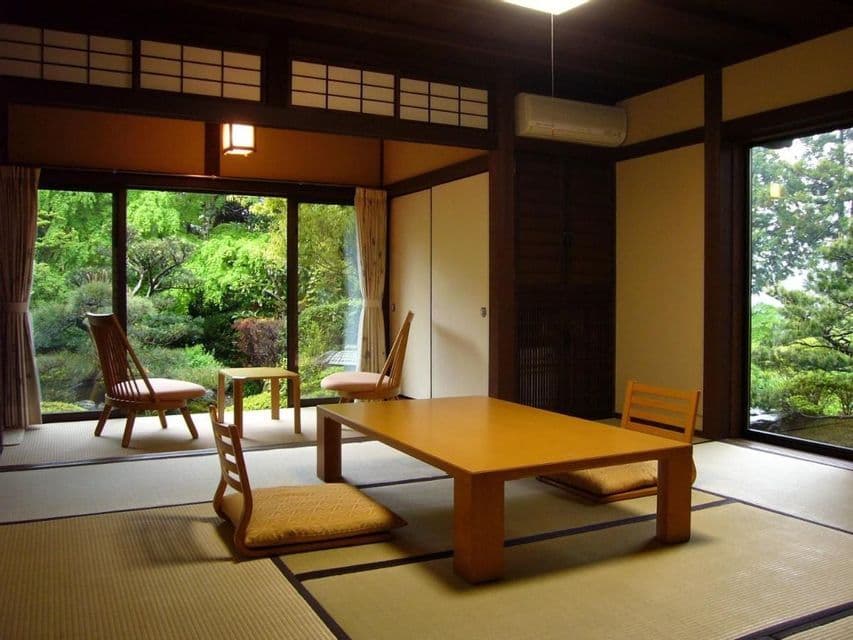 Una habitación japonesa tradicional con tatamis, una mesa baja de madera y sillas de suelo, con vistas a un exuberante jardín verde.