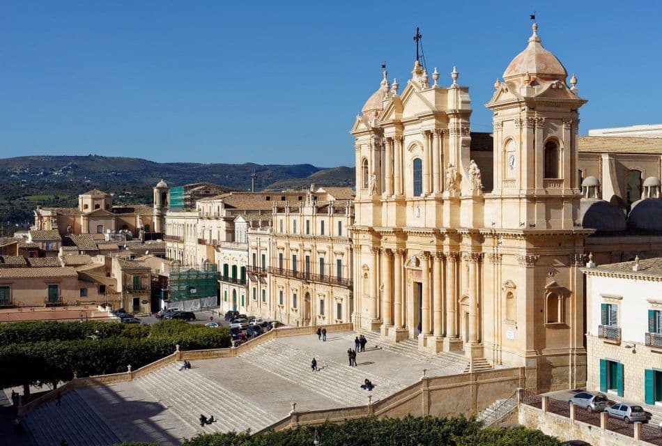 A sunlit, baroque-style cathedral with a wide staircase overlooks a historic town, with hills in the background.