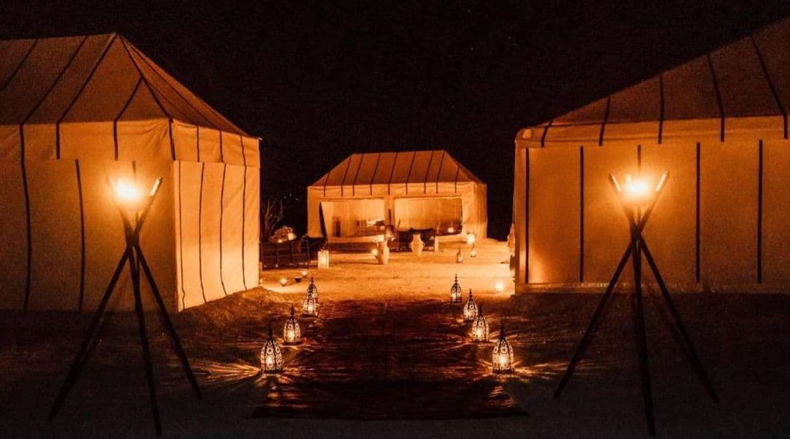 Des lanternes et des torches éclairent un chemin entre de grandes tentes de toile dans un camp du désert, la nuit.