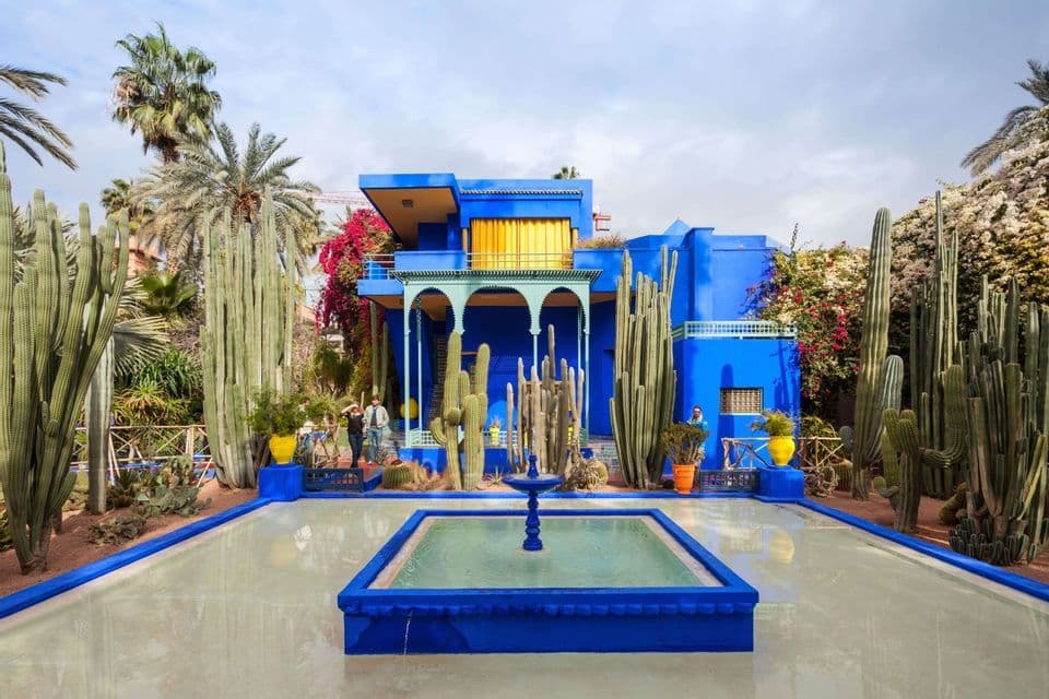 Un vivace edificio blu è immerso in un giardino di alti cactus e palme, con una vasca riflettente e una fontana di fronte.