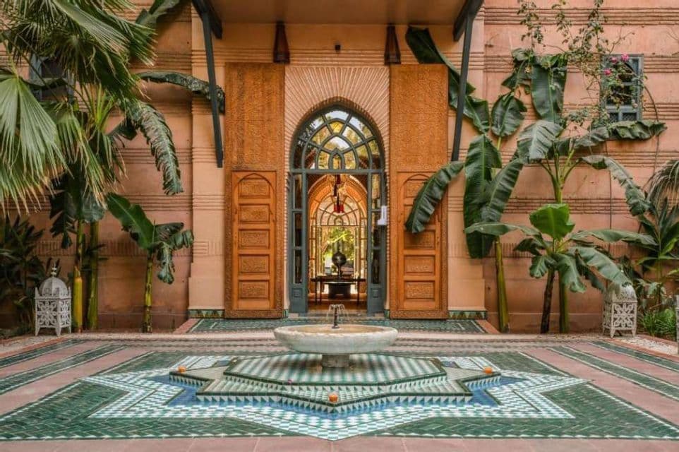 Una fuente en forma de estrella con azulejos de mosaico verdes y blancos se encuentra en un patio de estilo marroquí con una puerta arqueada y grandes plantas en macetas.