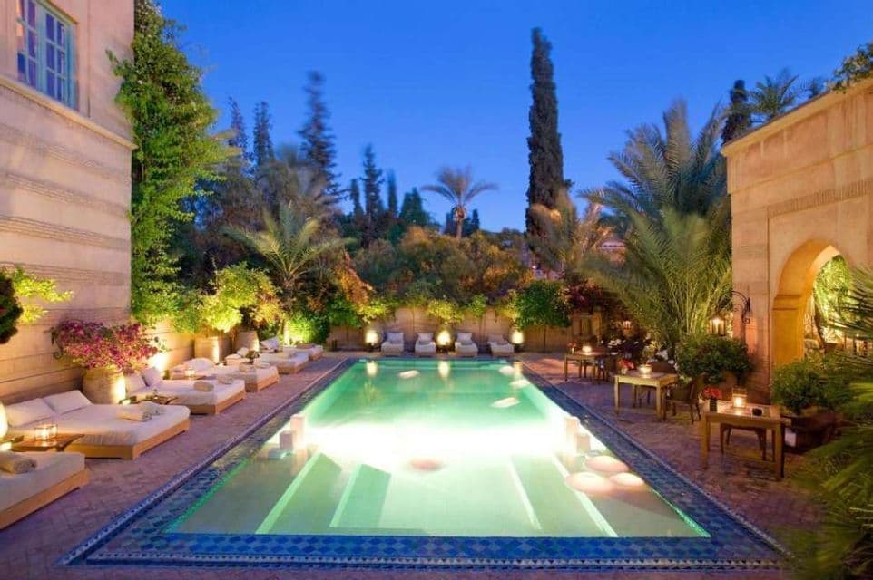 Una piscina rectangular iluminada en un patio al anochecer, rodeada de tumbonas y plantas exuberantes.