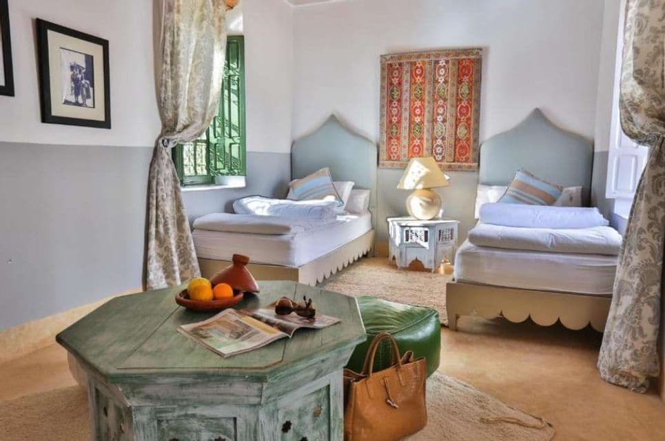 Une chambre avec deux lits simples, une table basse verte patinée et une décoration de style marocain, comprenant une tapisserie et un tajine.