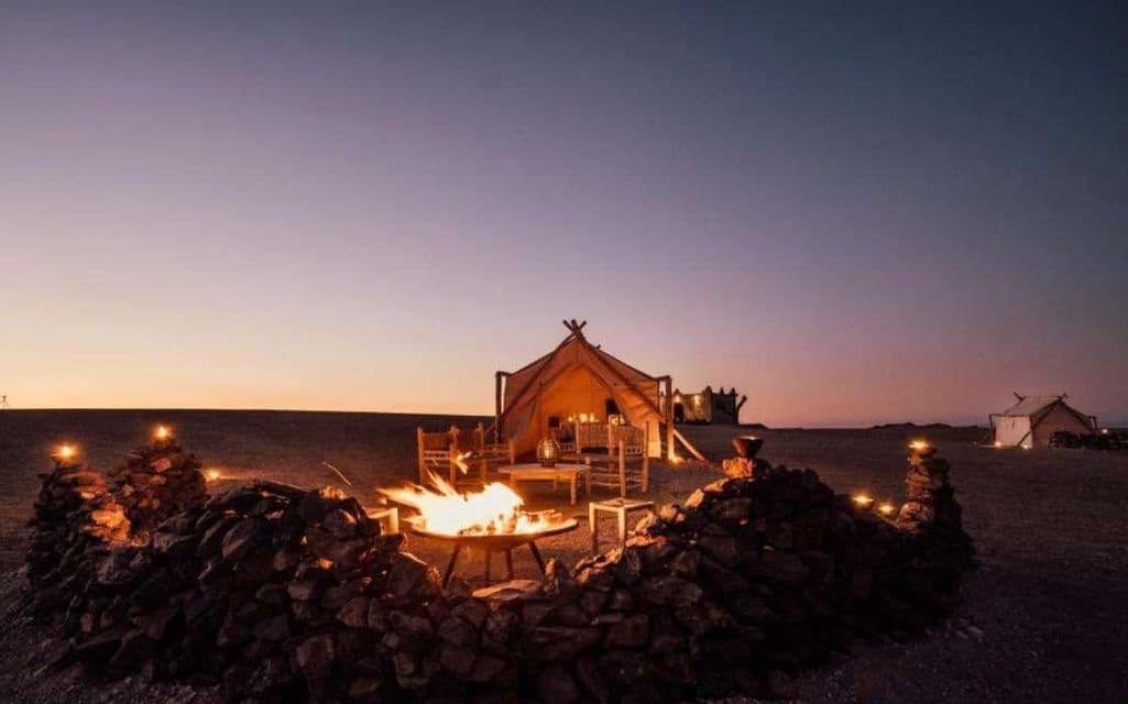 Un falò arde in una buca rivestita di pietre, davanti a una grande tenda illuminata in un campeggio nel deserto, al tramonto.