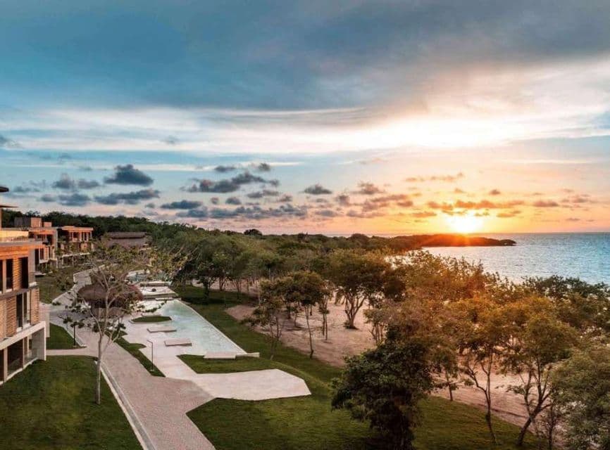Vue aérienne d'un complexe balnéaire avec des bâtiments modernes, des piscines et des arbres surplombant l'océan au coucher du soleil.