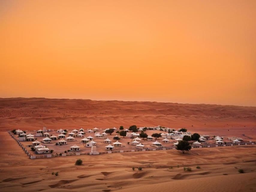 Ein weißes Zeltlager liegt in einem umzäunten Bereich inmitten sanfter Sanddünen unter einem orangefarbenen Himmel.