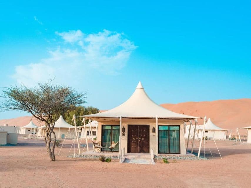 Une tente de glamping de luxe au toit pointu est installée dans un campement désertique, avec des dunes de sable en arrière-plan sous un ciel bleu.