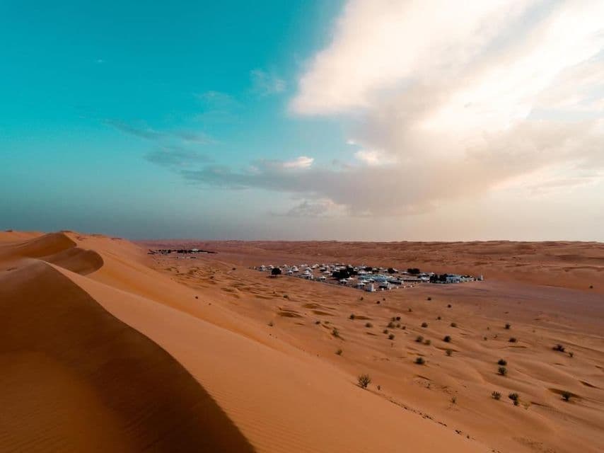 Eine Siedlung weißer Gebäude liegt eingebettet zwischen orangefarbenen Sanddünen in einer weiten Wüste unter einem türkisfarbenen Himmel mit Wolken.