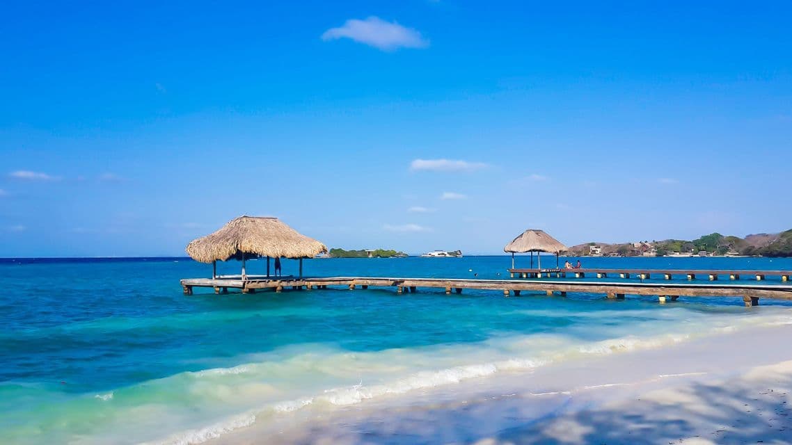 Une jetée en bois avec deux cabanes au toit de chaume s'étend sur l'eau turquoise de l'océan depuis une plage de sable blanc sous un ciel bleu clair.