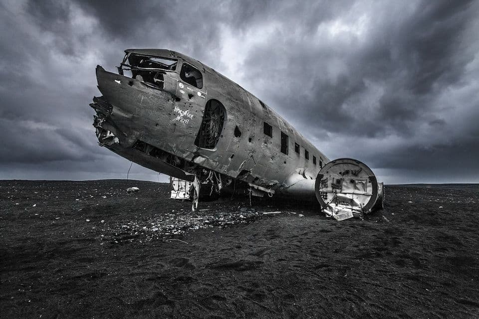 The wreckage of an airplane fuselage sits on a field of black sand under a heavily clouded sky.